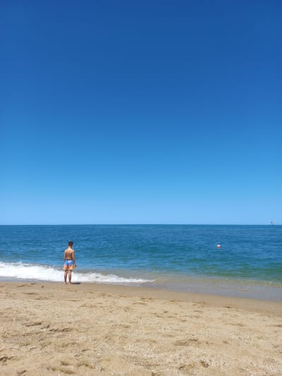 A young boy stands on a sandy beach, facing the calm blue ocean under a clear sky. The boy is wearing multi-colored swim trunks. Waves gently lap at the shore in front of him. Further out in the water, a small orange buoy and a distant boat are visible. The scene appears to be a serene day at the beach in Sinemorets, Bulgaria.
