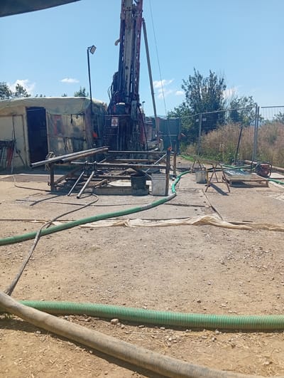 The image depicts an outdoor scene at a drilling site, likely in Chelopech, Bulgaria, given the context. A large drilling rig dominates the center of the frame, with its tall derrick and associated machinery. To the left, a temporary structure, possibly a shelter or tent, is visible, constructed from canvas or similar material and supported by metal poles. The ground is composed of dirt and gravel, with several pieces of equipment scattered around.
Prominently featured are multiple lengths of green, ribbed hoses snaking across the ground, suggesting water or fluid transfer is occurring. Metal pipes are also visible, some stacked and others connected to the drilling apparatus. In the background, a chain-link fence encloses the area, with vegetation and trees beyond. The sky is clear blue with a few wispy clouds.
The scene appears to be an active industrial or construction site. There are no people clearly visible in the image. The overall impression is one of a functional, albeit somewhat unkempt, work environment dedicated to drilling operations.