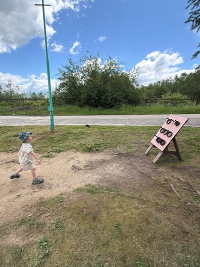 In the city of Kul'dur, Russia, a young child in a checkered outfit and a patterned hat is in mid-stride, about to throw a black ring. The ring is airborne, captured in motion. The child is on a dirt path, with dry grass and sparse green vegetation surrounding it. In the background, a road runs horizontally, bordered by more grass and dense green trees. Further beyond the trees, a chain-link fence and some indistinct structures are visible. A tall, green pole with a cross-shaped top stands on the left side of the image, near the child. To the right, a wooden target board stands on an easel, with several rings already placed on its pegs. The sky above is a vibrant blue, dotted with fluffy white clouds. The scene appears to be an outdoor recreational area.