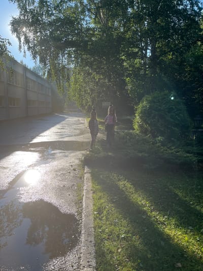 The image depicts a sunny outdoor scene, likely in Karagaiskii, Russia. The sun is bright, casting strong shadows and reflecting off a wet pavement or a large puddle in the foreground. The pavement is bordered by a grassy area on the right, and a line of trees and bushes on the left. In the background, a building with multiple windows and a corrugated metal facade is visible. Two individuals appear to be present in the scene, positioned between the trees and the grassy area. One person is wearing a pink shirt and dark pants, while the other is dressed in a checkered shirt and dark pants. Their presence suggests they might be walking or standing in this outdoor space. The overall impression is a bright, possibly humid day, with the sun's glare creating some lens flare and a hazy effect in certain areas of the image.