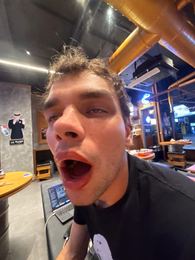 An extreme close-up, low-angle selfie, rotated roughly 90 degrees clockwise, captures a young man with light skin, dark, curly hair, and light-colored eyes. He wears a black t-shirt and has an exaggerated expression, with his mouth wide open, possibly mid-scream or shout, suggesting a playful or humorous mood.

The setting is an indoor commercial space, likely a bar or casual restaurant in Minhang District, China, characterized by an industrial-chic aesthetic. Prominent in the foreground are large, golden-colored ventilation ducts or pipes spanning the ceiling. Artificial, dim lighting suggests an evening or nighttime scene, with various light sources, including strip lights and distant neon signs casting a blue glow.

In the background, a black speaker with a blue indicator light is mounted high. On a dark table, an open laptop, likely a MacBook, displays text or code on its screen. A round wooden tabletop and a dark barrel or cylindrical object are also visible. A notable wall decoration features a stylized figure in a suit and fedora, reminiscent of a gangster, holding a bottle and a glass. Below this figure, Chinese characters are visible, though too blurry to decipher precisely. Distant signs in the background also display East Asian script. The overall atmosphere appears casual and lively.