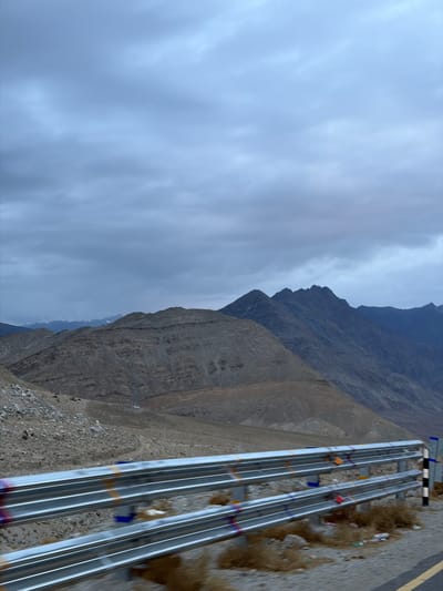 The media file depicts a mountainous landscape in Chumkar, India, under a cloudy sky. The scene is characterized by rugged, barren mountains with visible geological stratification. In the foreground, a road is visible, bordered by a series of metallic guardrails or pipes. These structures are somewhat reflective and appear to be painted with colorful ribbons or flags at intervals, suggesting a decorative or symbolic purpose, possibly related to local traditions or a significant route.
The ground next to the road is covered in gravel and sparse, dry vegetation. There are also some pieces of litter scattered along the roadside, indicating human presence. The mountains in the background are imposing and dominate the upper portion of the frame, with sharp ridges and varied textures of rock and soil. The sky is overcast, casting a diffused light over the entire scene, and hints at potentially cooler weather.
The perspective suggests the photo was taken from a slightly elevated position, looking down and across the landscape. The absence of people or a clear activity suggests a static scene, possibly a roadside view captured during transit. The overall mood is serene and somewhat stark, characteristic of high-altitude, remote environments. No visible text is present in the media file.