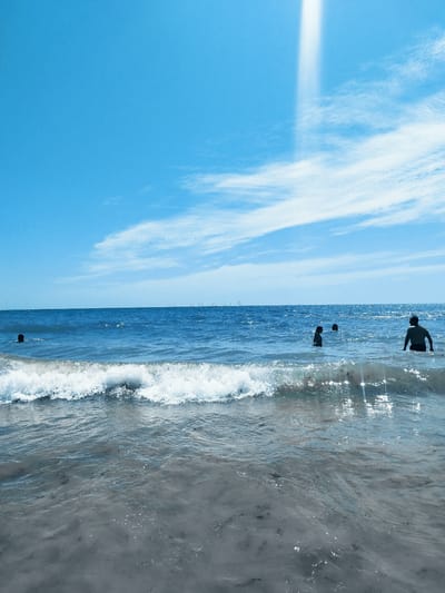 A wide shot captures a bright, sunny day at the beach in Rota, Spain. The clear blue sky dominates the upper half of the frame, with wispy white clouds scattered across it. A strong beam of sunlight shines down from the upper center, creating a bright, ethereal effect.

The lower half of the image is filled with the shimmering blue ocean. Gentle waves are rolling towards the shore, with a foamy white crest visible in the foreground. The water appears relatively shallow near the beach, with the sandy bottom visible beneath the surface in places.

Several people are enjoying the water. In the foreground, a wave breaks, obscuring some of the swimmers. Further out, in the middle ground, a group of four individuals are visible wading or swimming in the ocean. One person is on the left, with only their head and shoulders visible. To the right of them, two figures are closer together, and a third is further to the right, facing away from the camera.

In the far distance, on the horizon line, a fleet of small sailboats are scattered across the sea, adding a touch of activity to the vast expanse of water.

The overall mood is one of a pleasant summer day, with people enjoying a refreshing swim. The bright sunlight and clear skies suggest it is likely midday or early afternoon.
