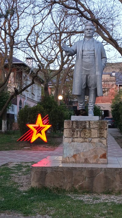The image shows a statue of a man in a long coat standing on a stone pedestal in what appears to be a park-like setting. A glowing red and yellow star with black and red stripes is positioned to the left of the statue. The statue is in Орджоникидзевский поселковый совет, Ukraine. A cat is sitting on the pedestal beneath the statue's feet. Behind the statue and star, there are buildings, trees without leaves, and a street light. A car is visible in the background to the right of the statue. The sky is a pale blue.