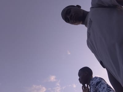 Two young men are present in the image against a light blue sky with scattered clouds. One man is positioned in the lower right of the frame, wearing a shirt with a floral pattern and resting his chin on his hand. The second man is positioned in the upper right, looming over the other, wearing a light-colored t-shirt. No evidence to ascertain the city or country can be obtained from the image.