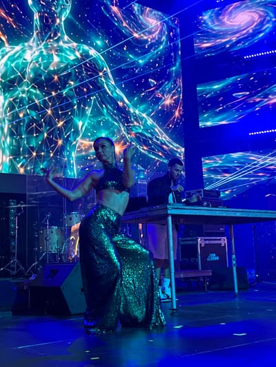 The image depicts a performance on a stage, possibly at an event in Akhalubani, Georgia, though this location cannot be confirmed from the visual information alone. A female dancer in the foreground is wearing a sparkling green sequined outfit, consisting of a crop top and wide-leg pants. She is in motion, with her arms extended and one leg bent. Behind her, a male DJ stands at a table equipped with electronic music equipment, including a Pioneer DJ setup. He appears to be operating the equipment. The stage is illuminated by vibrant blue lights, and a large digital screen in the background displays an abstract, cosmic visual of a human form interconnected with glowing lines and geometric patterns, alongside swirling nebula-like imagery. The environment suggests a concert, festival, or other live entertainment event.