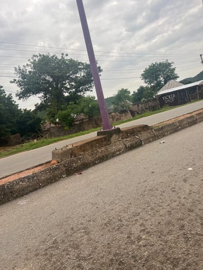 The image depicts an outdoor scene in Kona Garu, Nigeria, with a focus on a roadside and surrounding vegetation under an overcast sky. A paved road runs diagonally across the frame, bordered by a textured concrete curb. To the left of the road, lush green trees and foliage create a dense backdrop, with a few structures partially visible amidst the greenery. One building has a weathered, corrugated metal roof with visible text reading "TACKZA SHOP." A purple pole stands prominently in the foreground, extending vertically. Power lines crisscross the sky, suggesting an inhabited area. The overall mood is muted, likely due to the cloudy weather, which indicates it could be daytime but not particularly sunny. There are no people clearly visible in the frame, and the scene appears to be one of everyday life in a semi-rural or suburban setting.