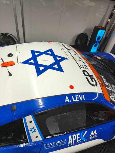 A racing car with a white, blue, and orange livery is situated indoors, likely within a garage or pit area, in Flatschach, Austria. The vehicle's surface is covered with water droplets. A prominent blue Star of David decal is centered on the white roof panel, accompanied by "GP ELITE" text, small red circular markers, a black antenna, and an orange antenna base. The driver's side features "A. LEVI" in white lettering on a blue stripe above the window. Below the window, a vertical stripe incorporates another blue Star of David. Further decals include "#CarreraCupDE" and sponsor branding such as "IDAN SHMUNIS INTERNATIONAL TAX SOLUTIONS," "APE MARKETING," and "MELAMED MOTORSPORT." The background consists of white walls, with a black floor fan visible to the left and an illuminated blue and black speaker unit beside a black fan to the right. The floor is dark and appears textured. No individuals are present.