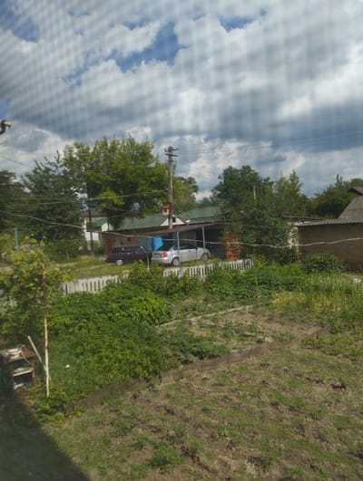 The image depicts a cloudy day in a rural or suburban setting, likely in Hnivan, Ukraine. The scene is dominated by an overcast sky with patches of blue peeking through. In the foreground, a patch of dry, tilled earth suggests a garden or agricultural area, with some small green plants growing. To the left, a makeshift shelf unit holds a white pot.
Further back, a white picket fence separates the garden area from a residential or utility space. Beyond the fence, two cars are parked under a covered structure attached to a building. One car is a dark-colored sedan, and the other is a lighter-colored hatchback. The building itself appears to be a garage or workshop, constructed with brick and a metal roof.
Behind the buildings, lush green trees and foliage provide a backdrop. Power lines are visible crisscrossing the sky, supported by utility poles, indicating the presence of infrastructure. The overall atmosphere is one of a somewhat unkempt but lived-in environment. The time of day appears to be daytime, given the brightness, but the cloudy conditions create a diffused light. No text is visible in the image.