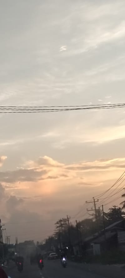 The image depicts a street scene in Keudeteupinpunti, Indonesia, during what appears to be late afternoon or early evening. The sky is overcast with a soft, diffused light, tinged with orange and pink hues, suggesting a sunset or sunrise. Power lines are visible horizontally across the upper portion of the frame, and more prominently in the mid-right section, connected to utility poles. The street below is lined with trees and buildings, some of which appear to be simple structures. Traffic is present, with several motorbikes and at least one car visible on the road. The motorbikes are in motion, indicating a busy street. The overall atmosphere is somewhat hazy, possibly due to dust or the low light conditions.