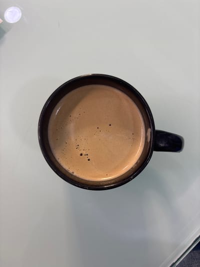An overhead shot depicts a black mug filled with a light brown beverage, likely coffee or a latte, sitting on a light-colored surface. The mug's handle is visible. Small bubbles or foam are present on the surface of the drink. The surface underneath is plain and light. The scene appears to be indoors. No people or landmarks are visible, so I cannot determine if it is located in Buenavista, Mexico.