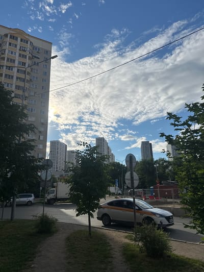 A bright, daytime scene in Kotelniki, Russia, features a residential high-rise building to the left, with multiple stories and balconies. A street lamp is visible in front of the building. In the middle ground, several modern, tall residential buildings stand against a sky filled with dynamic, white clouds. A taxi with orange and white markings drives on the right side of the road. A white truck is parked on the left side of the road. The foreground shows a dirt path and grassy areas with trees. Construction fencing and equipment are present in the background behind the taxi, indicating ongoing development.