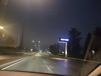 The image captures a nighttime scene on a highway in Laagri alevik, Estonia, viewed from inside a vehicle. A blue neon sign with the text "HEAD TIE'S!" is prominently displayed above the road. The highway is wet, indicating recent rain or damp conditions, and the road is illuminated by streetlights and the headlights of a car in the distance. Trees line the side of the road. The sky is dark and overcast.