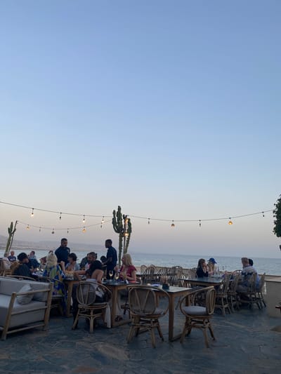 This image depicts an outdoor restaurant scene situated on a terrace overlooking the ocean, likely in Taghazout, Morocco, given the presence of surf-inspired décor and the coastal setting. The time appears to be late afternoon or early evening, as indicated by the soft, diffused light and the calm sea.
Several groups of people are seated at tables, engaged in dining and conversation. The architecture and furnishings suggest a relaxed, bohemian atmosphere, with rattan chairs and tables, and a light-colored sofa on the left. String lights with exposed bulbs are strung across the scene, adding to the ambiance. Two tall, sculptural cactus-like plants stand prominently, reinforcing the arid, coastal Moroccan aesthetic.
In the background, the vast expanse of the ocean stretches to the horizon, with a faint outline of land and buildings visible in the distance. Two men, presumably staff members, are visible standing and attending to patrons. The overall impression is one of a tranquil, upscale dining experience by the sea.