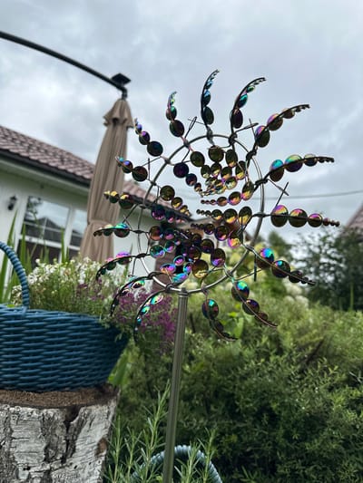 In the suburban garden of Ромашкино, Russia, a metallic wind spinner with iridescent, bead-like elements captures the viewer's attention. The spinner is positioned in the foreground, its reflective surfaces displaying a spectrum of colors against a backdrop of lush greenery and a partly cloudy sky. To the left, a blue woven basket filled with white and purple flowers sits atop a weathered birch log. In the background, the roofline of a house and a closed patio umbrella are visible. The overall scene depicts a tranquil outdoor setting.