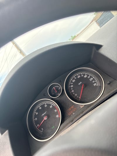 The media file displays the dashboard of a car, focusing on the instrument cluster. The scene is illuminated by bright daylight, with a clear sky and faint outlines of structures visible through the windshield. The dashboard itself is dark grey and shows some dust accumulation.
The main subjects are the car's gauges. A prominent speedometer, marked "km/h", shows a reading of 0 km/h, indicating the vehicle is stationary or moving very slowly. Its dial ranges from 0 to 260 km/h. To its right, a digital display shows the total odometer reading of "328429 km" and a trip meter reading of "16487". Below the speedometer, a smaller fuel gauge is visible, with markings "0", "1/2", and "1/1".
To the right of the fuel gauge, the tachometer is present, marked "x 100 min-1" and scaled from 0 to 80. Its needle also rests near 0. Several small indicator lights are visible around the gauges, including a green icon that resembles a headlight or parking light symbol. A portion of the steering wheel is visible on the left side of the frame. The overall setting is inside a car located in Karadjovo, Bulgaria.