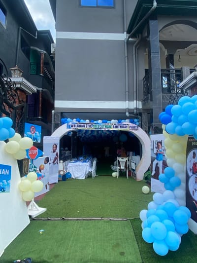 The image shows a birthday party entrance at a residence in Limbe, Cameroon, located at latitude 4.039 and longitude 9.202. The entrance is adorned with an archway decorated with blue and white balloons, and a banner that reads "Welcome to Joel Ethan Birthday Party." The archway leads to a pathway lined with white chairs, with a white table and blue and white balloons visible in the background. Several people are present in the background, some seated and some standing. The left side of the image features a sign that reads "Stop," and another sign that reads "It's Party Time." On the right side, there is a large cluster of blue and white balloons arranged in a cascading fashion. The ground in front of the archway is covered in green artificial grass.
