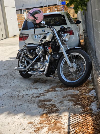 A black motorcycle is parked on a concrete surface next to a white car. The motorcycle has a black leather seat and chrome accents. A pink helmet with a black visor is attached to the back of the motorcycle. The car, a BMW, has a license plate that reads "BDJ 024". In the background, wooden crates are stacked and a grey chain-link fence runs along the right side of the image. Dry leaves are scattered on the ground. The scene appears to be an outdoor urban environment.