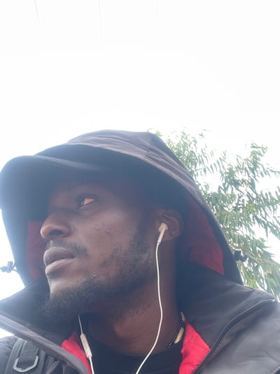 The media file is a close-up, low-angle selfie of a dark-skinned man wearing a black hooded jacket with a red lining. He has short, dark hair and a beard. He is wearing white earphones, with the wire visible across his chest. The man is looking downwards and to the left, with a neutral expression. The background consists of bright white sky and the leaves of a tree, suggesting an outdoor setting, possibly in Nigeria, given the location context. The lighting is bright, indicating it might be daytime. The image is oriented sideways.