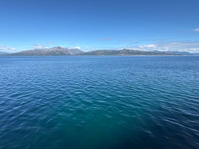 This is a landscape photograph taken from the water, looking towards a mountainous coastline under a clear blue sky. The foreground is dominated by the rippling surface of the ocean, with varying shades of blue and turquoise indicating depth. Gentle waves create a textured pattern across the water.
In the background, a long, rugged mountain range stretches across the horizon. The mountains appear barren and rocky, with some patches of snow or lighter-colored rock visible on their peaks. The coastline is irregular, with inlets and promontories. The sky is a vibrant, cloudless blue, with a few wispy white clouds scattered high up near the horizon.
The scene suggests a bright, sunny day, likely during the daytime. The clarity of the sky and the calm water indicate fair weather. The presence of mountains and a large body of water points to a coastal or fjord region. Given the provided location context, this is likely a view of the Norwegian coast, specifically Lødingen, Norway, characterized by its dramatic mountainous scenery meeting the sea. There are no visible people or specific objects that draw attention, allowing the focus to remain on the natural landscape. The overall mood is serene and majestic, emphasizing the grandeur of nature. No text is visible in the image.