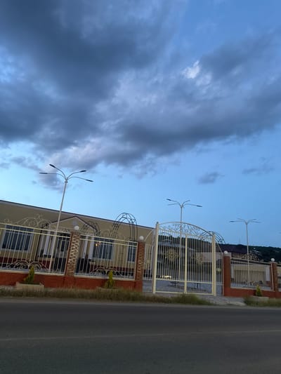 The image depicts a roadside scene in Manavi, Georgia, under an overcast sky at dusk or dawn. The upper two-thirds of the frame are dominated by a sky featuring light blue patches towards the right and bottom-center, transitioning to a pervasive, heavy coverage of dark grey stratocumulus clouds across the upper left and central portions.
In the mid-ground, a substantial property is visible, enclosed by an ornate white metal fence and gates. The fence features decorative scrollwork and vertical pickets, supported by pillars with a reddish-brown brick-like facade and topped with white spherical light fixtures. A prominent, large white metal gate, matching the fence's intricate design, provides vehicular access in the center-right. To the left, a cream or light yellow building facade with reddish-brown lower walls is visible behind the fence, featuring multiple window-like openings with white railings. Above this building, a roofline with a central pediment-like element is discernible. Further architectural elements, including arched metal structures, are integrated into the fence line. Several unlit, modern streetlights with four radiating arms are positioned along the street, two prominent on the left and one on the right, extending above the property's height.
The foreground is occupied by a dark asphalt road. Between the road and the property fence, a strip of dry, sparse yellowish-brown grass and earth is present, punctuated by two small, conical green shrubs planted in grey rectangular planters. In the distant background, behind the property, a gentle hillside with sparse vegetation and additional building structures is partially visible under the twilight sky. No people or vehicles are present in the scene.