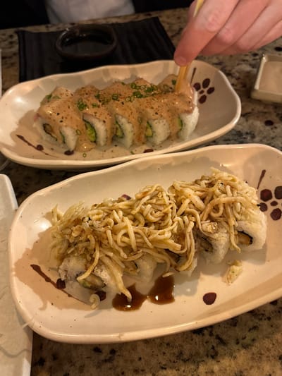In Magdalena del Mar, Peru, two plates of sushi are presented on a speckled countertop. The foreground plate holds four sushi rolls topped with crispy, shredded ingredients and a drizzle of sauce. The background plate displays six sushi rolls covered in a creamy sauce and garnished with chopped green onions. A hand, holding chopsticks, is reaching towards a sushi roll on the background plate. A small dish of dark liquid, likely soy sauce, is positioned behind the background plate. The lighting is dim, suggesting an indoor setting.