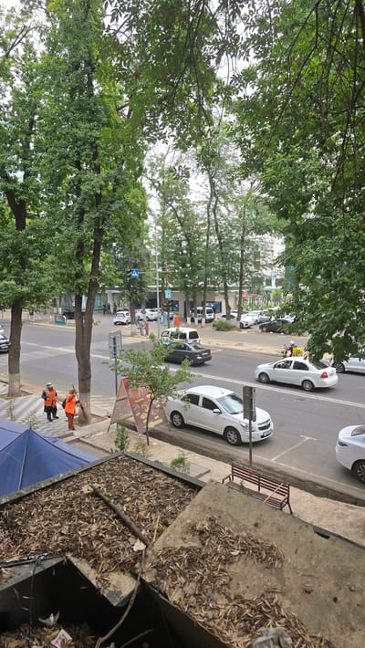 In the city of Katta Chilanzar-2 MFY, Uzbekistan, a street scene unfolds with vehicles and pedestrians. Two sanitation workers, one in an orange vest and black pants, the other in an orange vest and trousers, are visible on the sidewalk near a blue awning. The worker on the left appears to be observing, while the worker on the right is sweeping. Across the street, cars are driving and parked, including a white sedan and a dark sedan. Several people are walking near a crosswalk in the distance. Trees line the street, with buildings visible in the background. The foreground is dominated by a close-up view of a rooftop covered in debris and leaves, with a metal rod and part of a broken structure. A red park bench with a broomstick leaning against it is situated on the sidewalk further down the street. The sky is overcast.