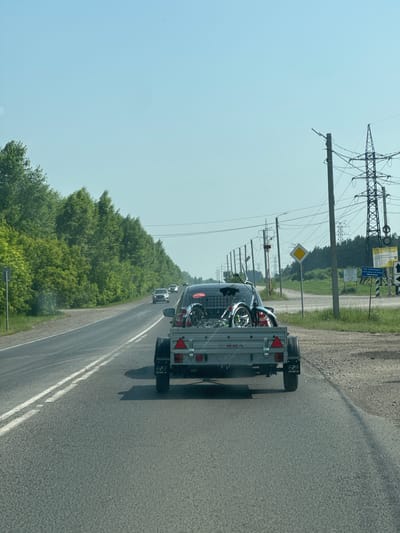 The image shows a view from inside a vehicle traveling on a paved road lined with trees. The road has a white double line marking the center. In front of the vehicle is a trailer being towed by a car, carrying two bicycles. Several other cars are visible on the road ahead. To the right of the road, there are utility poles with power lines and various signs. The sky is clear and blue. The environment suggests a rural or suburban setting. The sign on the trailer reads "МЗСА".