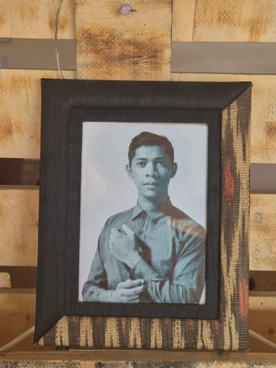 A framed, black and woven photo of a young man with short, dark hair and wearing a button-down shirt is hanging on a wooden pallet. The portrait is slightly rotated, showing the man looking at the camera. The woven pattern on the outer frame is tan with brown and red stripes. A purple pushpin and electrical wire are visible on the pallet to the left of the frame. The background is primarily composed of the wooden pallet structure. It is not possible to determine the location within Pasay, Philippines from this image.