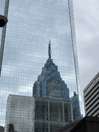 The image shows a low-angle view of a glass skyscraper in Philadelphia, USA, reflecting another building. The reflection appears distorted due to the irregular surface of the glass facade. The sky is overcast, casting a gray tone over the scene. A security camera is visible in the lower left corner. Part of another building with a striped exterior is seen in the top. Another building with a glass exterior and some windows can be seen to the right of the reflective building.