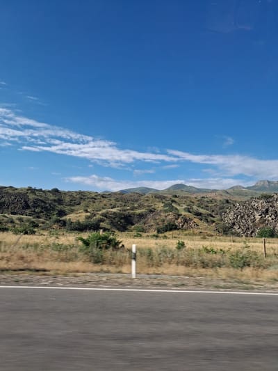 A horizontal, eye-level, medium-shot photograph captures a scenic landscape from a moving vehicle, as indicated by the motion blur on the foreground asphalt road. The road, a dark gray asphalt surface with a thin white line marking its edge, occupies the bottom quarter of the frame. Beyond the road, dry, golden grass with sparse green shrubs stretches across the mid-ground. A single white and black striped pole stands upright in the field, partially obscured by the motion blur. The background features rolling hills covered in low-lying green vegetation and rocky outcrops, characteristic of the semi-arid terrain found in Malishka, Armenia. The sky above is a vibrant, clear blue, dotted with scattered white clouds that cast subtle shadows on the landscape. The overall impression is one of a vast, open natural environment under a bright, sunny sky.