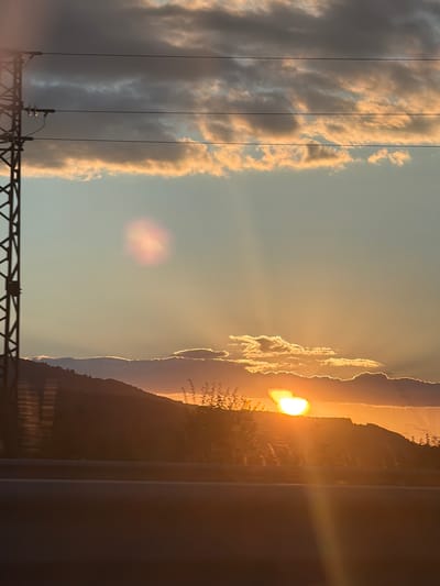 This image depicts a sunset over a mountainous landscape in or near Studena, Bulgaria. In the foreground, a dark, indistinct barrier, possibly a guardrail or a road surface, runs horizontally. Behind this, a dark silhouette of hills and sparse vegetation is visible against the bright sky. The sun is partially obscured by clouds and the crest of a hill, casting strong rays and a warm, orange glow. The sky is a gradient of pale blue and orange, with dramatic clouds illuminated by the setting sun, appearing in shades of gray, yellow, and pink. A power line tower with multiple wires stretches vertically on the left side of the frame, partially obscuring the sky and clouds. A faint lens flare is visible in the upper left quadrant of the image. The overall impression is of a natural landscape at dusk, with man-made electrical infrastructure present.