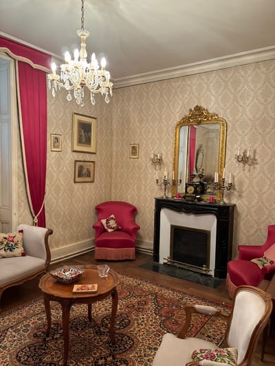 This media file captures an opulent, classically decorated interior room, likely a living room or salon, in Étréchy, France. The scene is static, with no activity or people present.

The room features walls covered in a light-colored wallpaper with a subtle, damask-like pattern. A grand crystal chandelier with multiple lit bulbs hangs from the ceiling, casting light across the space. Below it, rich, deep red curtains with decorative trim are draped to one side, hinting at a window or doorway.

Dominating one wall is a large, ornate gold-framed mirror positioned above a black and white marble fireplace. The mantelpiece holds a decorative clock and a pair of candelabras. On either side of the mirror, gold wall sconces contribute to the room's illumination.

Several pieces of antique-style furniture are arranged around the room. Two plush armchairs, upholstered in a vibrant magenta or deep pink, are visible; one has a fringed edge and an embroidered cushion. A light-colored settee or sofa with wooden accents and an embroidered cushion is also present. Between some of the seating, a small, round wooden side table with carved legs holds a decorative dish and a clear ashtray. The floor is largely covered by a magnificent oriental rug featuring intricate floral and medallion patterns in shades of red, blue, and gold, set upon what appears to be a hardwood floor. A few framed pictures of varying sizes adorn the walls, adding to the room's traditional decor.

The artificial lighting from the chandelier and sconces suggests an indoor setting, possibly in the evening or a dimly lit space. There is no visible text in the image. The overall impression is one of historical elegance and comfort.