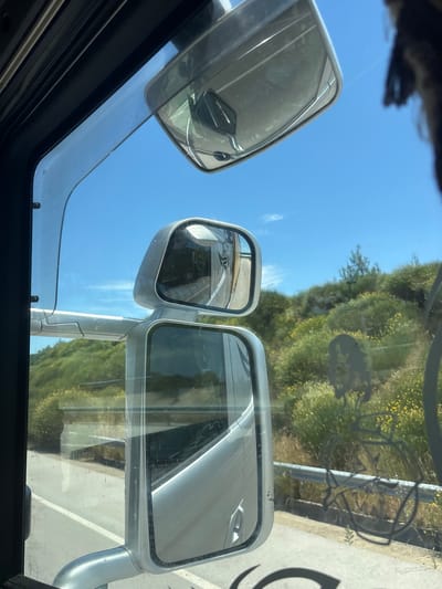 The image is taken from inside a vehicle, looking outwards through the side window and mirrors. The viewpoint suggests the vehicle is in motion, likely a truck or bus, given the size of the mirrors. The scene depicts a bright, sunny day with a clear blue sky and scattered white clouds. Outside the vehicle, a green, hilly landscape is visible, covered with vegetation including bushes and small trees. There are yellow wildflowers scattered throughout the greenery. A road with a white line is visible, bordered by a metal guardrail. The mirrors reflect the surrounding environment, including parts of the vehicle itself and the landscape. There is a distinct shadow on the window that appears to be a stencil of a bicycle. No people are visible, and the image focuses on the exterior environment as seen through the vehicle's windows and mirrors. The location is stated to be Λύρα, Greece, suggesting a rural or semi-rural setting within that region.