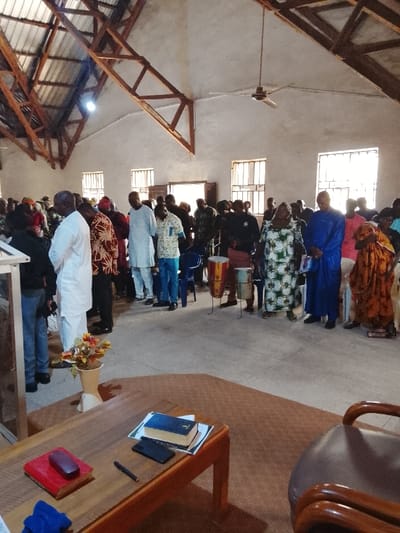 The image shows a gathering of people inside a building with a wooden roof. The room appears to be a church or a place of worship. It has a tiled floor and several windows on the walls. There are about 20 people present, both men and women, dressed in casual clothing, some standing and some seated. The people appear to be interacting and engaging in a religious activity. There are two drums in the middle of the room, along with a microphone stand and a small wooden table. There are flowers in a vase next to the table. A large wooden table sits in the foreground, with a bible and other items on it. A chair is visible in the lower right corner. The coordinates provided are not a location in Mukaiya, Nigeria, but rather in the Atlantic Ocean. The image depicts an indoor scene in a church setting.
