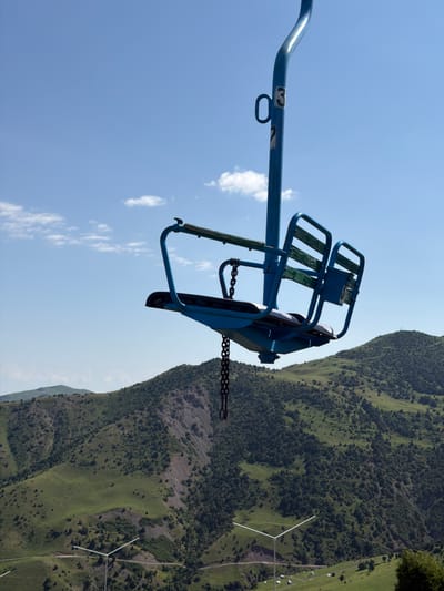 A blue chairlift seat is suspended in the air against a bright blue sky with scattered white clouds. The chairlift seat is empty and appears to be made of metal with wooden slats for the seating and backrest. A black chain hangs from the bottom of the seat. The chairlift post is also blue and has the number "3" and "7" visible on it. In the background, rolling green hills covered in trees and shrubs stretch out under the sky. A road is visible winding through the hills. There are also some white structures that appear to be wind turbines or similar equipment on the lower slopes of the hills. The scene is a view of a mountain landscape, likely in a recreational area.
