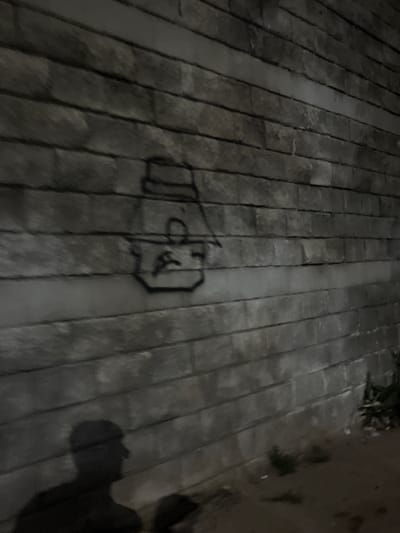 A shadowy, low-light image captured at night shows a rough-textured, grey brick wall with horizontal mortar lines. A crude black graffiti drawing of a face wearing a hat is present on the upper portion of the wall. In the lower left, a dark silhouette of a person's head and upper body is cast on the wall, suggesting a light source behind the viewer. The ground in front of the wall appears to be dirt or sand, with some sparse, dark vegetation visible on the right. The overall scene is gritty and urban.