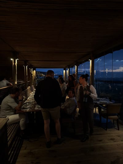 A group of people gather around a long table at a restaurant with a scenic view. A server in a white shirt and vest pours a beverage. Diners are casually dressed, some in collared shirts. The restaurant's decor features wood, with a woven ceiling and wooden floors. It's dusk, with a view of lights from a city across the water.
