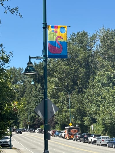 The image depicts a street scene in Port Moody, Canada, likely on a sunny day given the clear blue sky and bright lighting. A dark green lamppost stands prominently in the foreground, displaying a colorful banner with a stylized pink flamingo against a backdrop of yellow, blue, and green abstract shapes. Attached to the lamppost is a dark green streetlamp with a white bulb. A diamond-shaped traffic sign is also affixed to the lamppost.
The street, appearing to be a multi-lane road, is lined with parked cars and a food truck adorned with flames. Trees with lush green foliage line the sides of the road, creating a verdant environment. In the distance, more vehicles and trees are visible. The overall impression is a suburban or semi-urban setting with a focus on street infrastructure and natural surroundings.