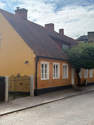 The image depicts a narrow cobblestone street in Lund, Sweden, with a two-story building dominating the scene. The building features an orange-yellow facade and a red-tiled roof with multiple chimneys. Several white-framed windows are visible on the building's facade. A black wrought-iron gate with decorative circular elements is positioned in front of a portion of the building, bordered by white posts and a wooden fence. A large, leafy tree grows near the side of the building, partially obscuring the entrance. The sky above is cloudy, with a mix of blue sky and white clouds. The street surface is made of evenly laid cobblestones, with a raised concrete curb running along its edge. The overall impression is of a historic European streetscape.