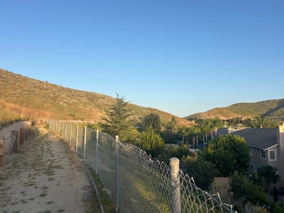 The image shows an outdoor scene in Oak Park, United States of America. A dirt path runs parallel to a chain-link fence, with wooden posts and rope on the left side. Beyond the fence are residential houses partially obscured by trees and shrubs. In the background, there are rolling hills covered in dry vegetation, with a clear blue sky above. The lighting suggests it is either early morning or late afternoon. There are no people visible in the image.