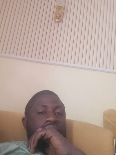 A single adult male, appearing of African descent, is depicted in a medium-shot selfie, looking directly into the camera. His right hand, adorned with a ring featuring a red stone on a finger, rests near his chin. He is wearing a light-colored, possibly green, shirt. To his right, a segment of a yellowish-brown upholstered armchair or sofa is visible. The immediate background consists of a plain, light beige wall. Above the wall, a white paneled ceiling is present, with a gold-toned light fixture or sensor, featuring a central glass element, mounted on the panels in the upper right quadrant. The scene is indoors. The specified location in Babawa, Nigeria, cannot be visually confirmed from the image itself.