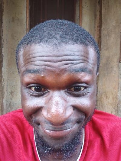 This is a close-up, rotated, eye-level shot of a Black man's face. He is wearing a red t-shirt with a white trim around the neckline. The background appears to be a rough, wooden surface, possibly a wall or floor, with horizontal planks. The lighting suggests it is daytime, but there are no strong indicators of specific weather conditions. The man has short, dark, curly hair and a light beard. His eyes are open, and he is looking directly at the camera. His expression appears neutral or perhaps slightly relaxed. The image is a portrait focusing on the man's facial features, with a shallow depth of field that blurs the background.