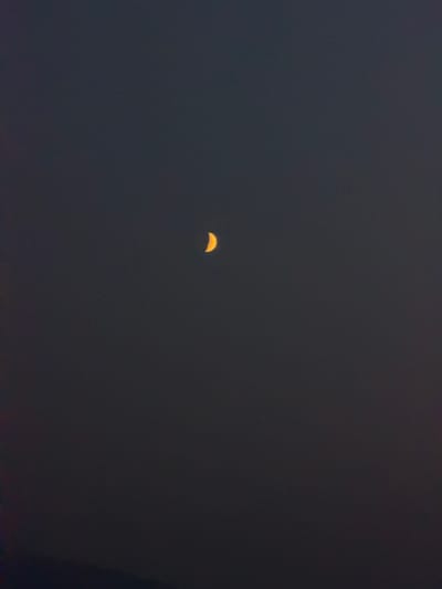 The image shows a crescent moon in a dark, overcast sky. The moon is small and appears yellow-orange. The sky is a uniform dark gray, with some subtle variations in tone. There is a hint of land at the very bottom of the image, suggesting it might be a night sky view from a terrestrial location. The image does not contain any information about the city of Усть-Мана, Russia, or any people, objects, or actions.