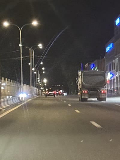 The image depicts a nighttime scene of a road, likely a highway, in Tairov, Armenia. The perspective is from a vehicle traveling on the road, looking towards the side. The foreground shows a streetlamp with multiple illuminated bulbs, casting a warm glow. Further down the road, a line of similar streetlamps stretches into the distance, creating a sense of depth. The road surface appears wet, reflecting the lights and suggesting recent rain or damp conditions.
On the left side of the road, there are signs of infrastructure, possibly overpasses or bridges, with some blue and red lights visible on them. A large vehicle, likely a truck, is partially visible on the overpass. The overall atmosphere is dark and urban, characteristic of a city at night. The lighting creates a moody and somewhat abstract effect. No people are clearly discernible in the image. The activity taking place is simply travel on a road at night. The time of day is clearly nighttime, and the wet road surface hints at possible weather conditions. Location cues are limited to the name "Tairov, Armenia." There is no visible text in the image.
