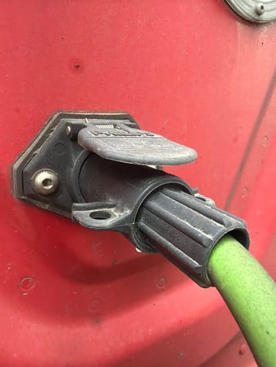 The image shows a close-up, eye-level view of a black electrical connector socket mounted on a red, textured surface, likely the exterior of a vehicle or piece of equipment. A green hose or cable is inserted into the connector. The socket is hexagonal with a hinged cover, which is partially open, revealing the internal contacts. A silver screw is visible on the left side of the socket, securing it to the red surface. The surface itself has several small, circular indentations and some dust or debris. The lighting suggests an outdoor environment, and the overall impression is one of functional, utilitarian equipment. There are no people or other distinguishing features in the image. The provided information does not specify a location in Murfreesboro, United States of America.