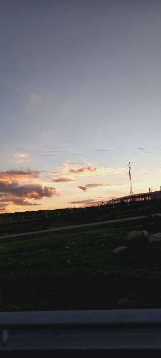 A serene landscape unfolds, featuring a colorful sky at dusk, transitioning from dark gray to warm hues of orange and pink. Fluffy clouds are scattered, some tinged with hints of red. A transmission tower stands against the backdrop. Patches of green ground cover the foreground. A vertical structural element occupies the extreme right edge of the frame.