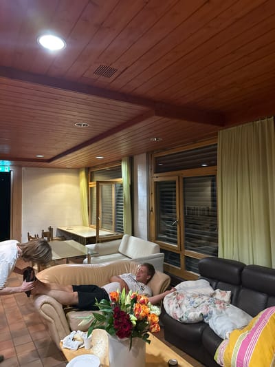 This image depicts an indoor scene, likely a common area or lounge, within a building located in Wald (ZH), Switzerland. The room features a prominent wooden-paneled ceiling with recessed lighting fixtures. On the left side of the frame, an emergency exit sign with green lettering and a running figure is visible.

The central focus of the image is a man reclining on a light-colored leather sofa, wearing a white t-shirt and black shorts. He is facing towards the right, with his legs extended. Another person, partially visible on the left, is bending down and appears to be interacting with the reclining man's feet, possibly removing or adjusting a shoe. This individual is wearing a white t-shirt.

In the foreground, a wooden coffee table holds a bouquet of colorful flowers, primarily roses in shades of red, orange, and yellow. There are also plates, a white cup, and some crumpled napkins on the table.

To the right of the reclining man, a dark brown leather sofa is visible, covered with floral-patterned bedding and cushions. A portion of a striped blanket or pillow in yellow, pink, and green is also visible on this sofa.

In the background, a large window with wooden frames and venetian blinds is present, partially obscured by pale yellow curtains. Adjacent to the window, a white brick wall is visible. Further in the background, there are tables and chairs, suggesting a dining or seating area. The floor is tiled with reddish-brown square tiles. The overall lighting suggests an evening or indoor ambiance.