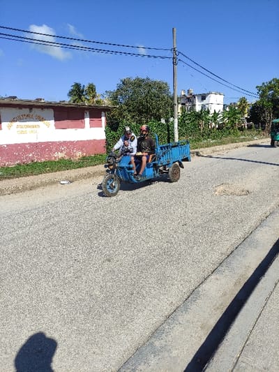 The scene depicts an outdoor urban environment in Ciego de Ávila, Cuba, under a clear blue sky with sparse white clouds. A blue three-wheeled motorized vehicle, designed with a small cargo bed at the rear, travels on an asphalt road. The road exhibits visible wear, including cracks and a prominent pothole in the mid-ground. Two adult males are aboard the vehicle; the driver wears a white long-sleeved shirt, dark pants, and a light-colored helmet, with hands on the handlebars. The passenger, seated directly behind, wears a dark t-shirt, dark shorts, and a dark helmet with red accents. To the left of the road, a single-story building with a red base and white upper section displays partially visible yellow text reading "AGRICULTURA CIEGO DE AVILA." Green vegetation lines the space between the building and the road. Multiple black utility wires are strung across the sky. On the right, a utility pole stands amidst dense green foliage and trees, behind which several light-colored multi-story buildings, one with a rooftop water tank, are visible. A portion of a green three-wheeled vehicle is discernible in the far right background, partially obscured by trees. Bright sunlight illuminates the scene, casting shadows in the foreground.