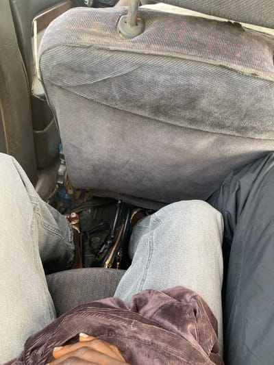 The image depicts the interior of a vehicle in Unguwar Gaya, Nigeria, viewed from a high-angle perspective. In the immediate foreground, a person's dark-skinned hands, with manicured fingernails, are clasped and resting on a dark brown, ribbed fabric garment (possibly corduroy). Light gray denim jeans are visible on this person's lap. To the right, a portion of a leg clad in a dark gray or black fabric garment is visible.
In the midground, the worn backrest of a gray fabric vehicle seat occupies the upper half of the frame. The fabric is discolored and shows signs of wear, with a lighter, slightly patterned border at the top edge. A metal headrest support post extends upwards from the seat. Below the seat and between the legs of the foreground person, a black handbag with a visible gold-colored zipper and decorative elements is placed on the floor. Adjacent to the handbag, a person's feet, wearing open-toed sandals adorned with small, reflective embellishments, are visible. One foot displays painted toenails. These feet are connected to legs clothed in light gray denim jeans, consistent with the person occupying the seat in front of the viewer. The vehicle's interior trim, possibly a dark gray door panel, is visible on the far left. The overall environment suggests a compact and well-used vehicle cabin.